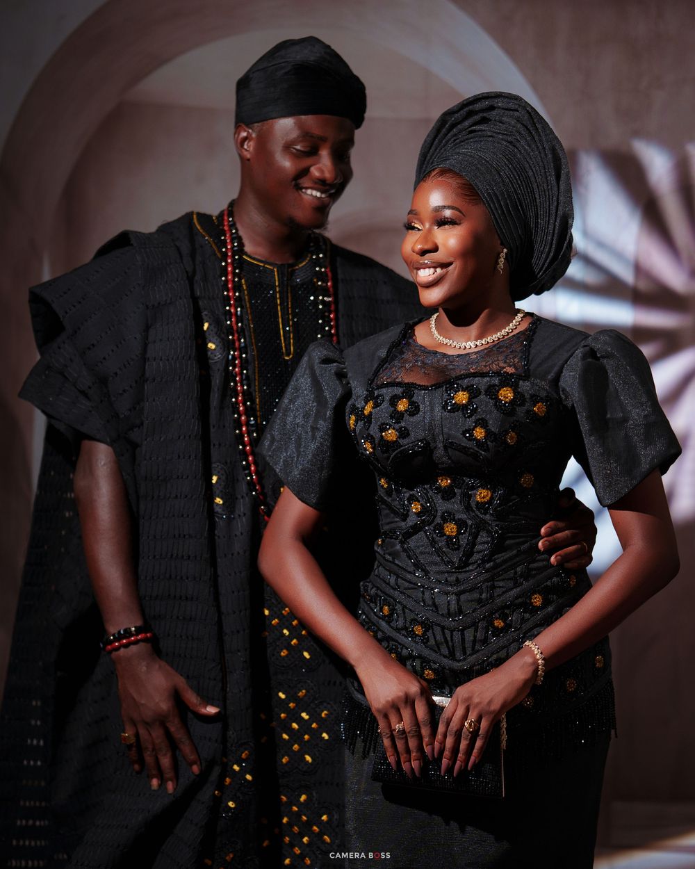 Nigerian couple in matching black traditional aso-oke outfits with gele and cap smiling together.