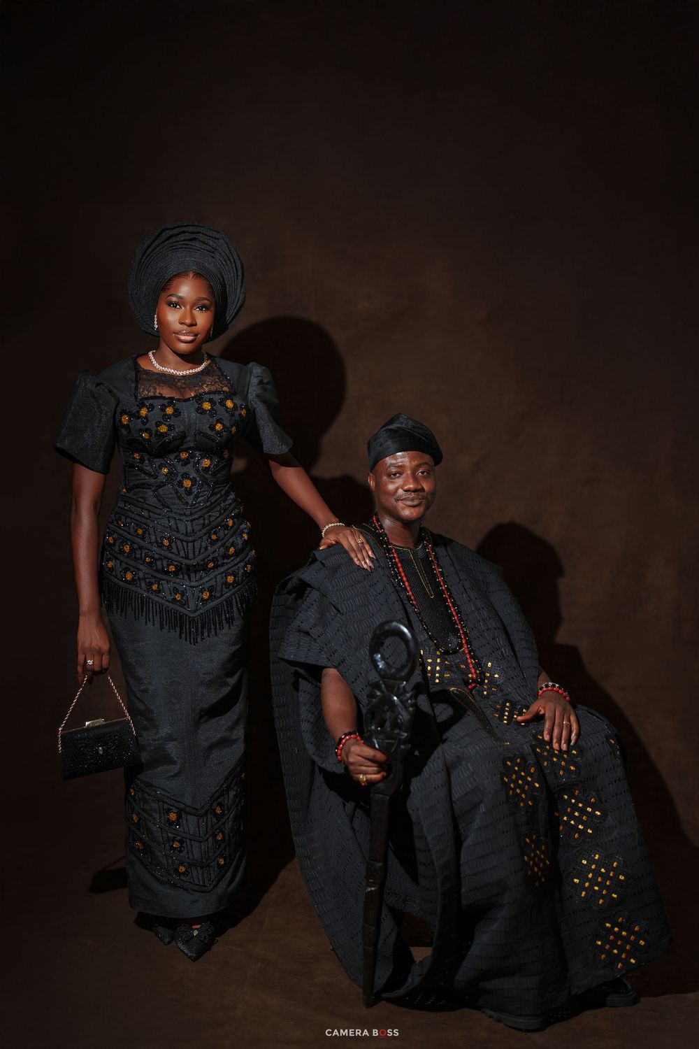 Two people in matching dark traditional African attire pose elegantly for a formal portrait photo.