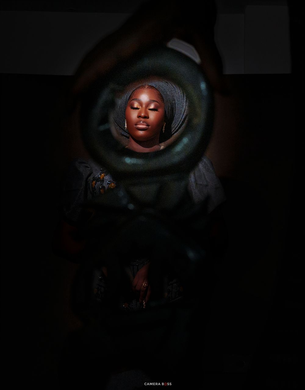 A young Black woman in a head wrap poses serenely, framed by dark hands forming a circle around her illuminated face.