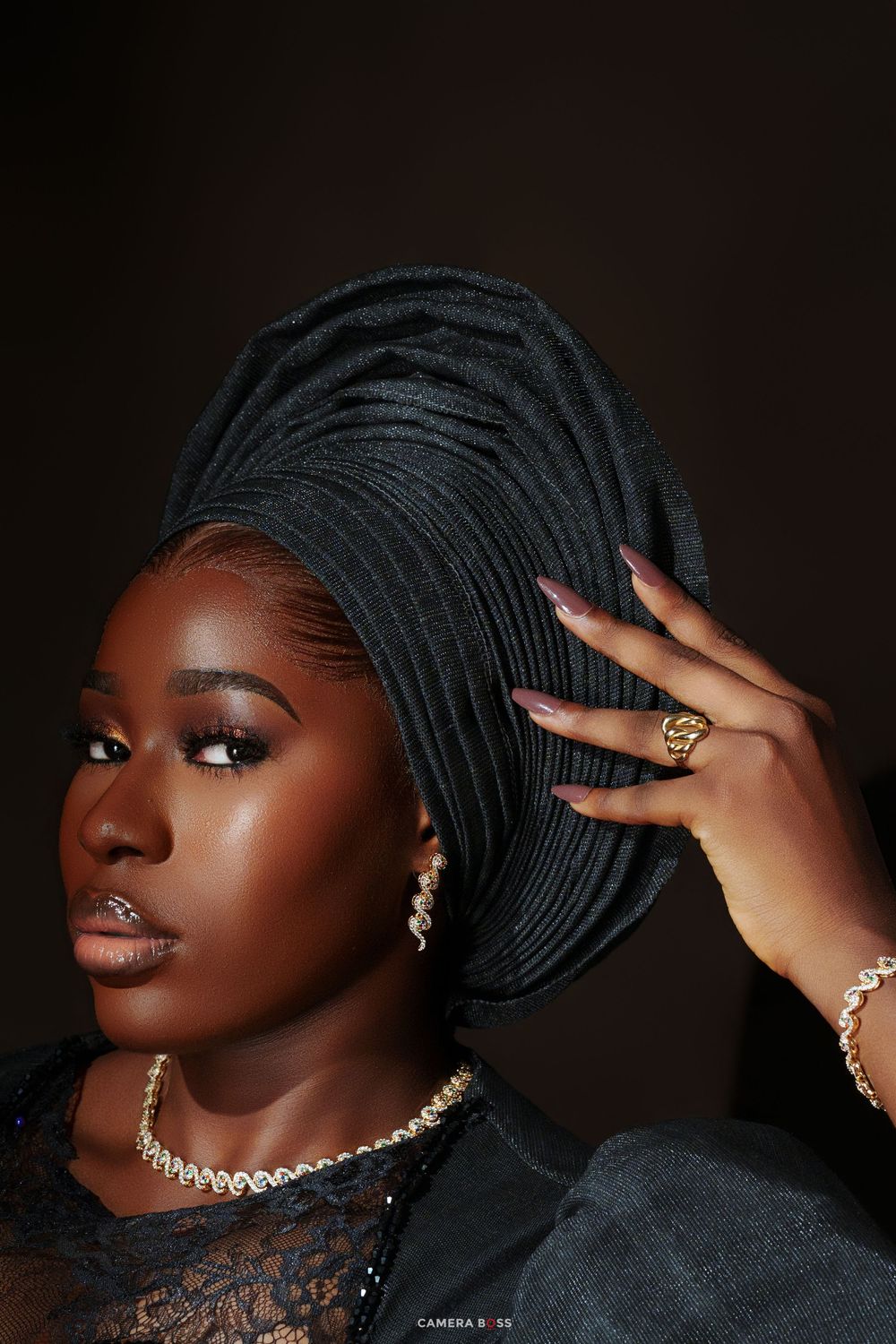 Close-up portrait of woman wearing black gele headwrap with gold jewelry and lace outfit.