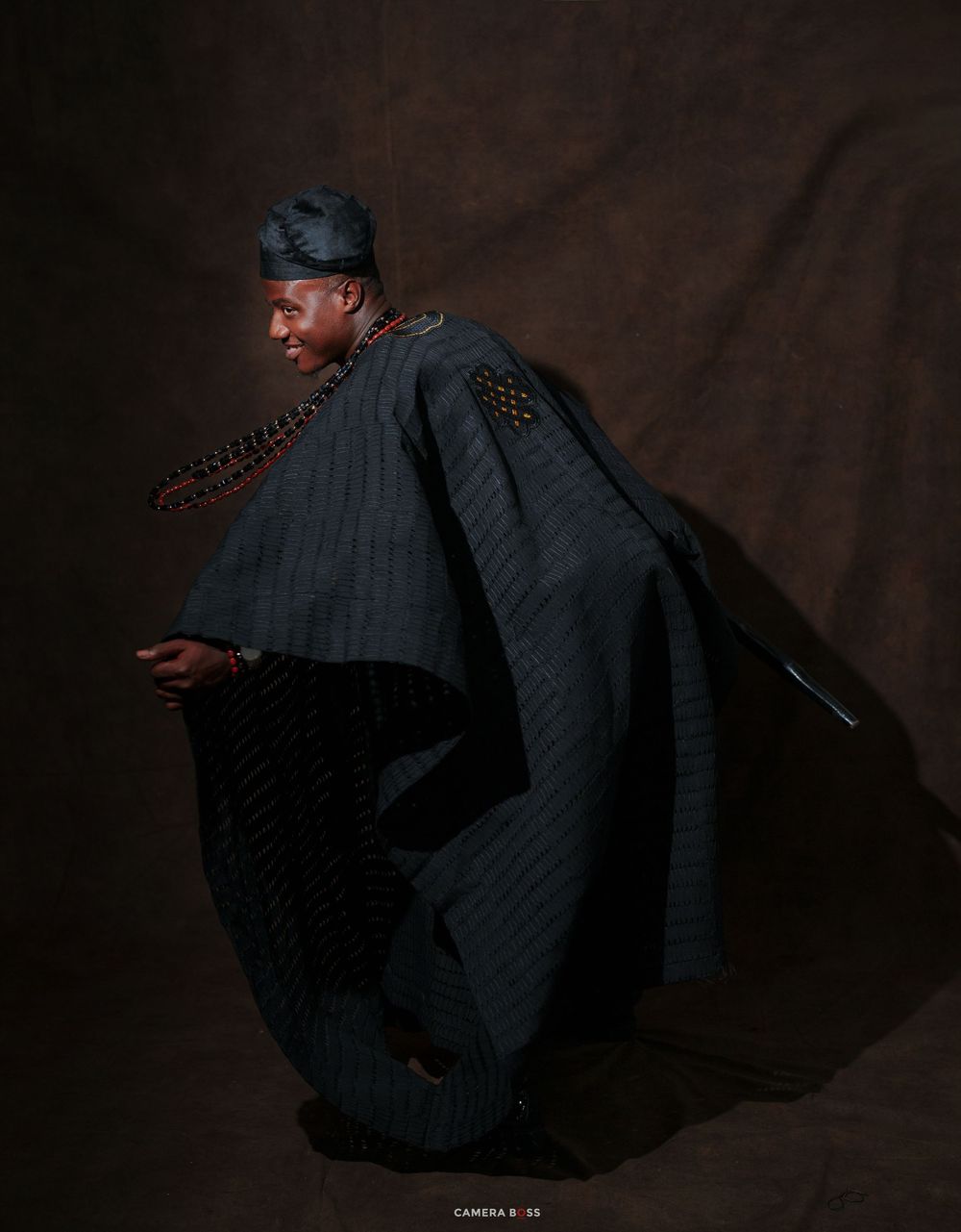 Man in traditional African agbada attire with beaded necklace and matching cap poses elegantly against dark background.