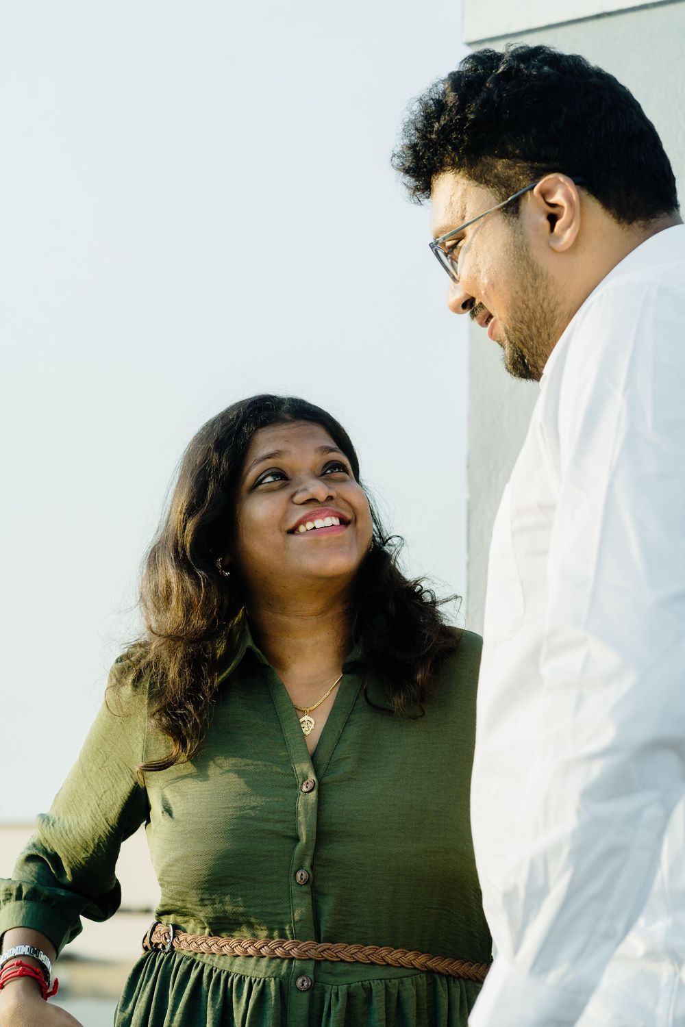 Smiling Indian couple gazing at each other outdoors, woman in green dress, man in white shirt with glasses.