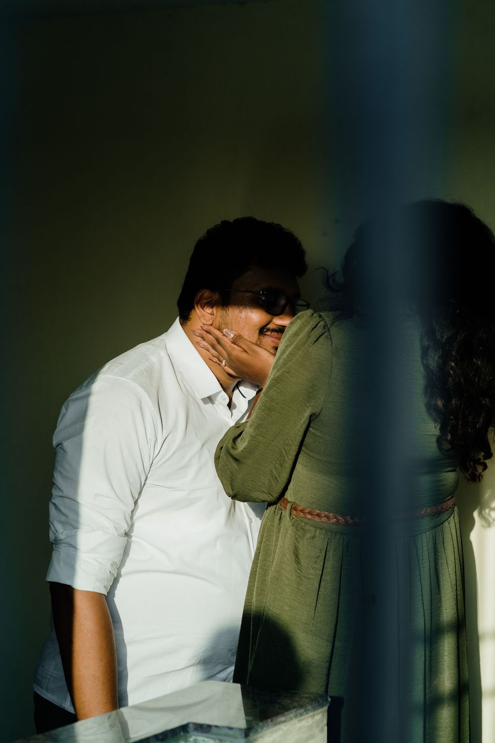 A man in a white shirt tenderly holds a woman's face while sharing an intimate moment against a sunlit wall.