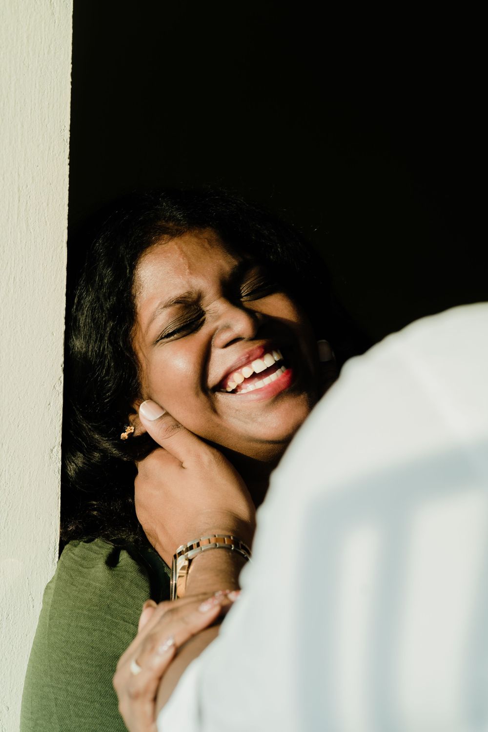A joyful South Asian woman laughs warmly while leaning against a sunlit white wall, wearing a green top and gold bracelet.
