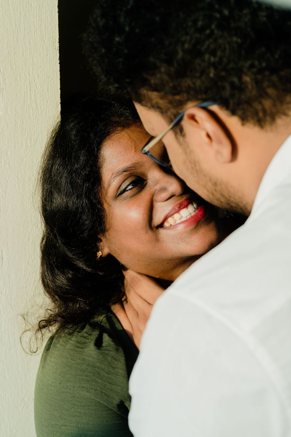 A smiling South Asian woman in a green top being embraced by a man wearing glasses against a wall.