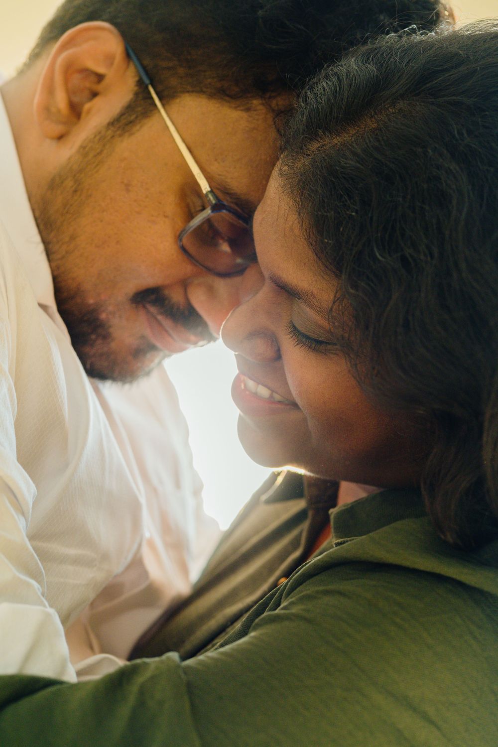 An interracial couple shares an intimate forehead-to-forehead moment, smiling tenderly at each other indoors.