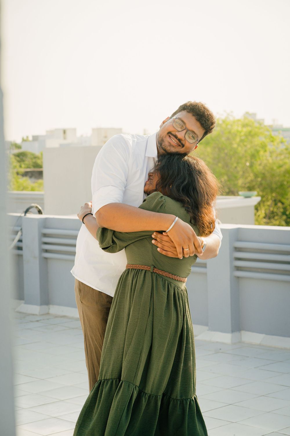 A happy South Asian couple shares a warm hug on a rooftop terrace, man in white shirt smiling over womans shoulder.