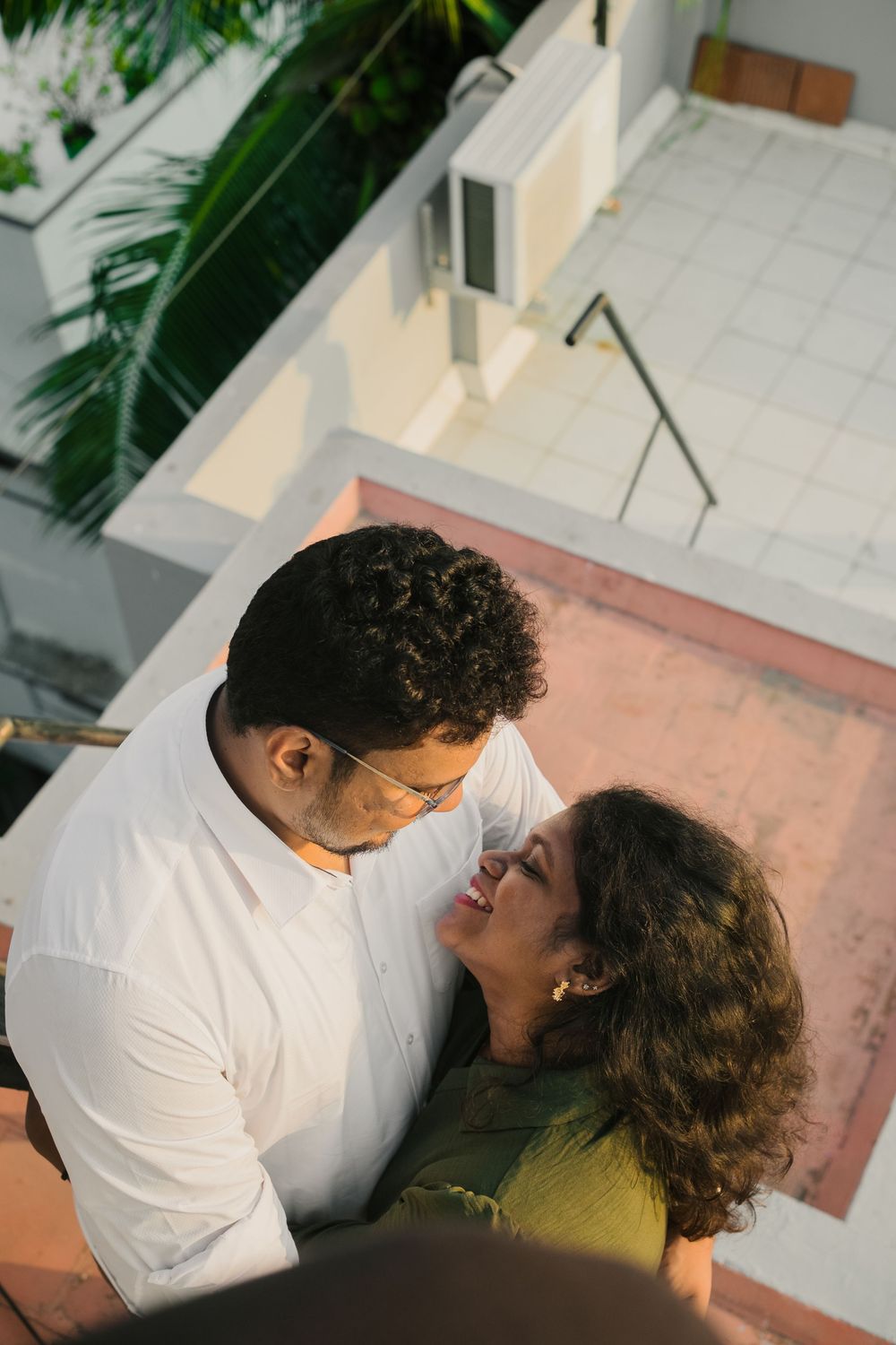 A romantic couple sharing an intimate moment on a balcony, smiling at each other with lush greenery in the background.