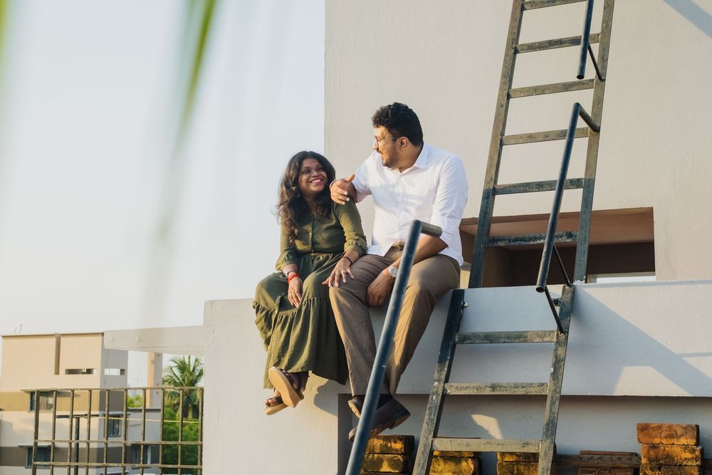 A couple sits together on a rooftop ledge beside a metal ladder, smiling and enjoying a relaxed moment outdoors.