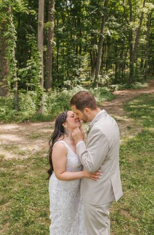 Bride and groom share a romantic moment in a lush forest setting, with the groom in a tan suit and bride in a lace gown.
