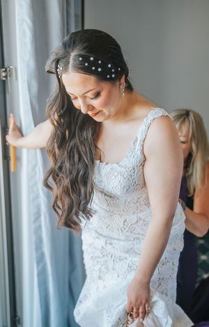Bride in lace wedding gown with pearl hair accessories, smiling as bridesmaid helps with dress near window.