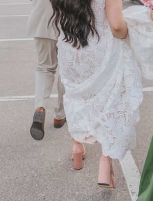 Bride in white lace dress and pink block heels walking with groom in tan suit on pavement.