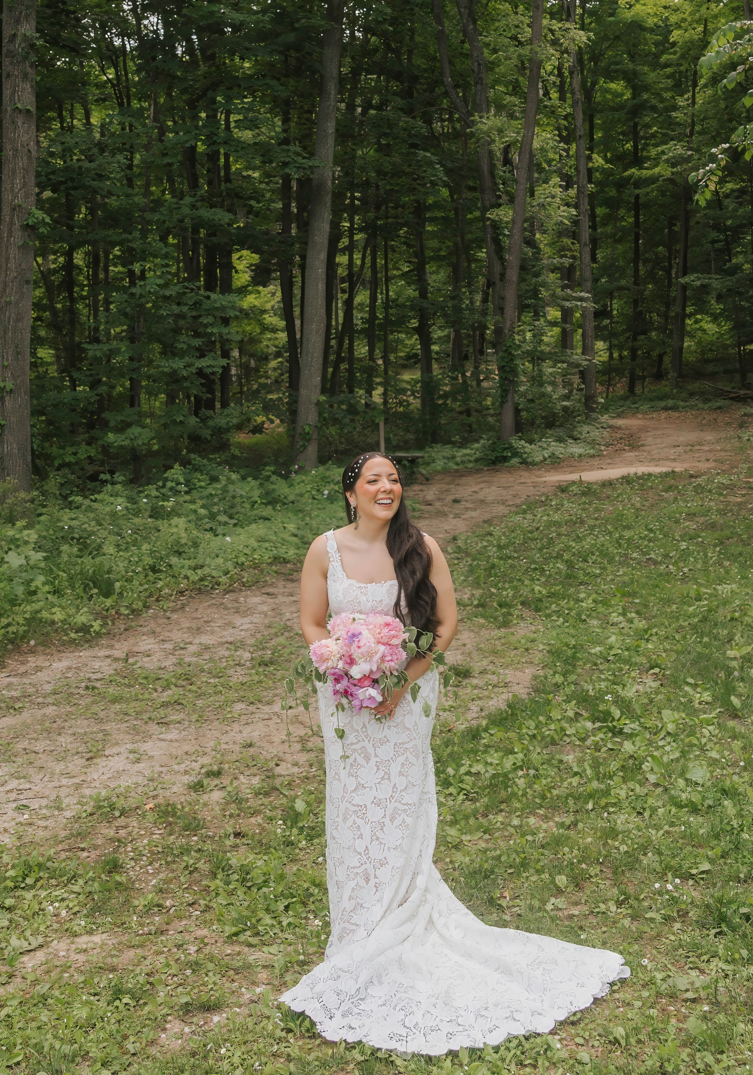 Smiling bride in lace wedding gown holding pink bouquet standing on forest path surrounded by lush green trees.