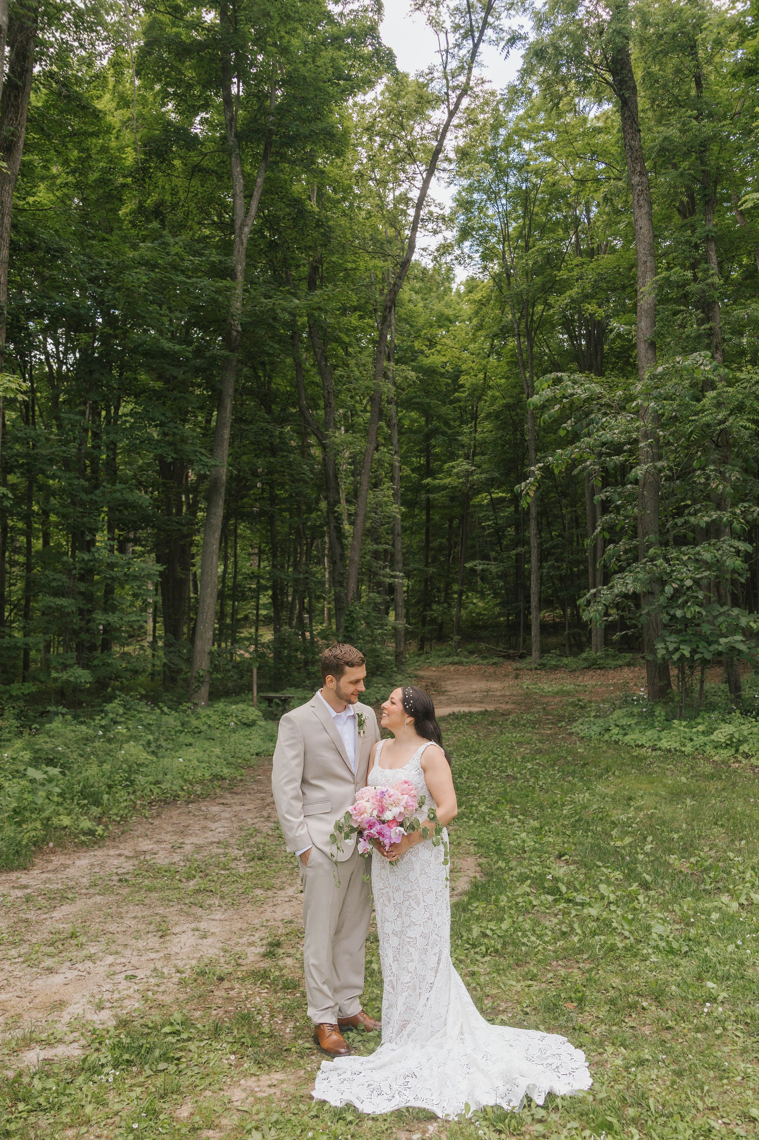 Bride and groom standing together in a lush green forest clearing, bride holding pink bouquet in lace wedding gown.
