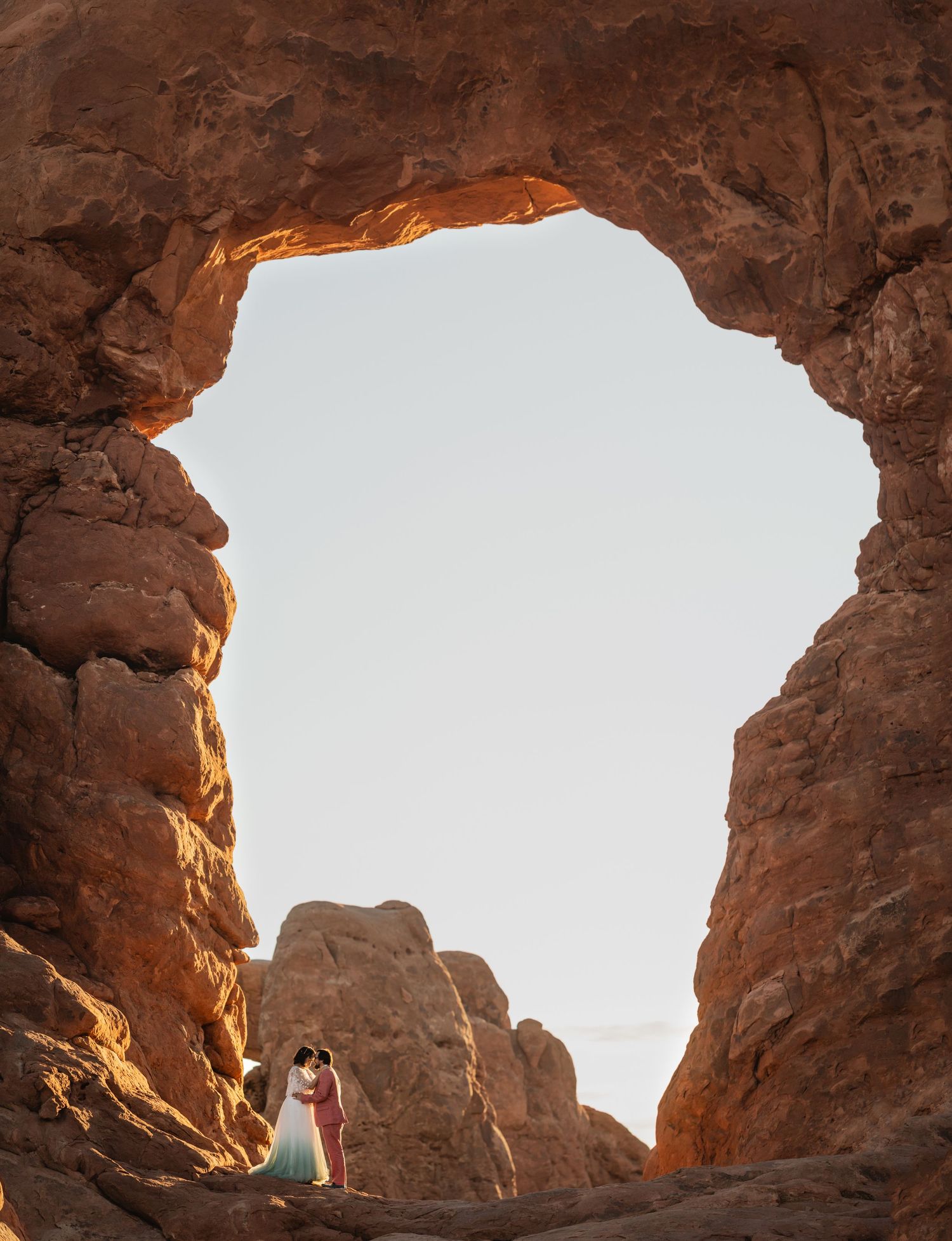 Wedding couple shares intimate moment framed by dramatic red rock arch at Arches National Park.