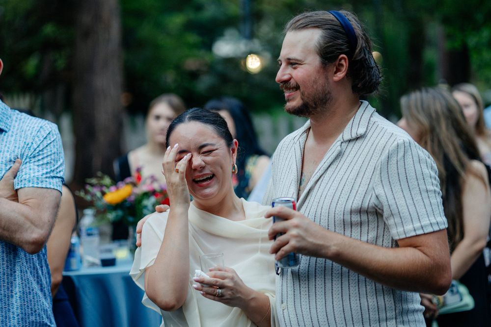 Guests laughing and crying with joy at an outdoor evening wedding reception surrounded by trees and string lights.