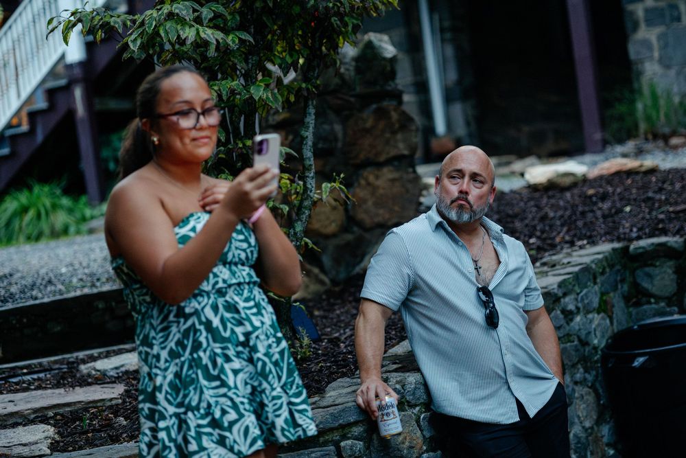 Woman in floral dress photographing with phone beside bald man holding drink at welcome dinner at the Blind Tiger hotel in Asheville.