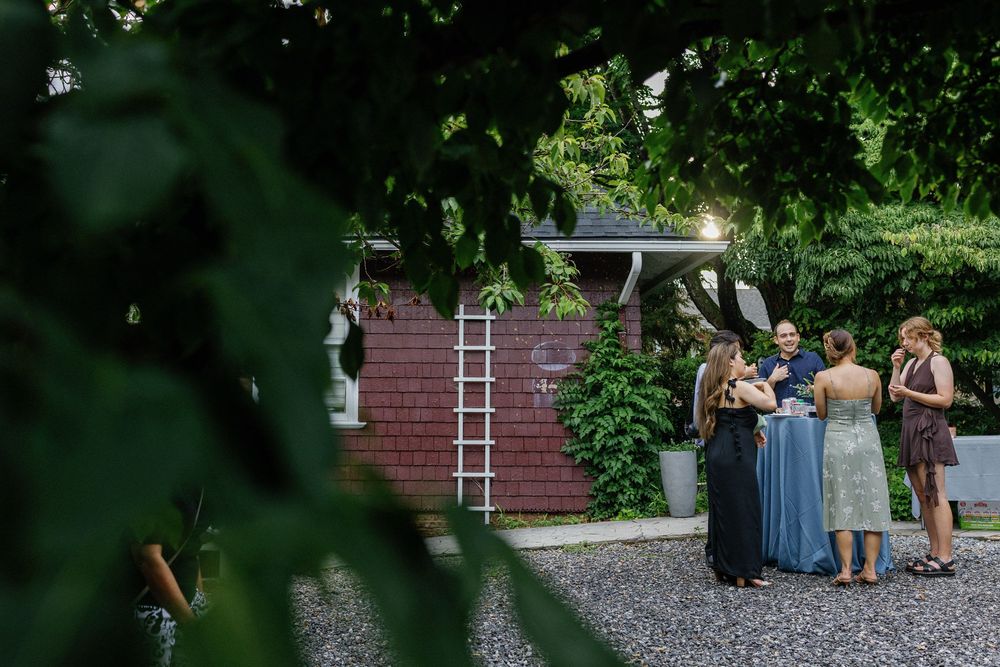Wedding guests mingling outdoors near a red brick shed surrounded by lush greenery at dusk.