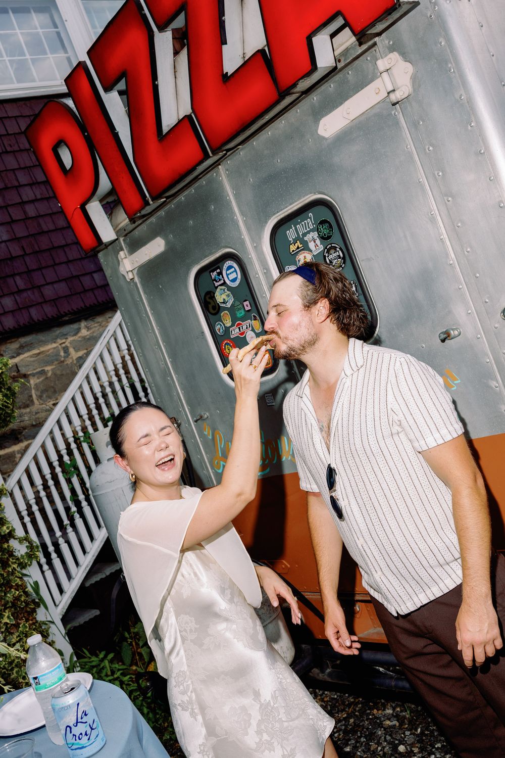 Bride laughing as she feeds groom pizza from a food truck at their outdoor wedding reception.
