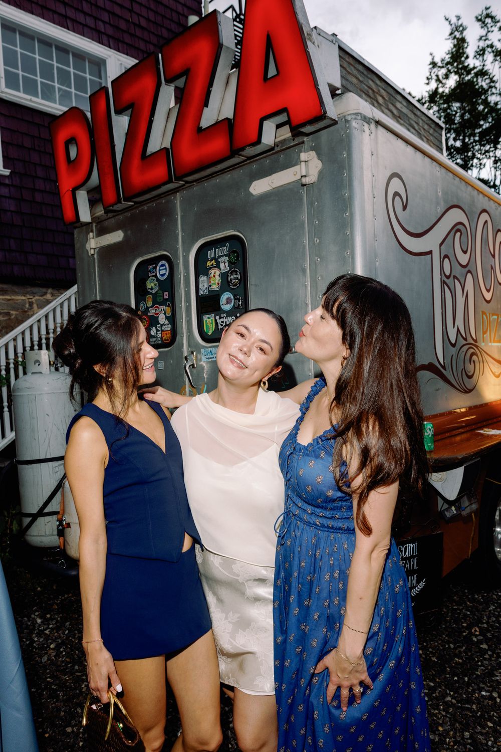 Three women in elegant dresses smile and embrace in front of a pizza food truck at an outdoor evening event.
