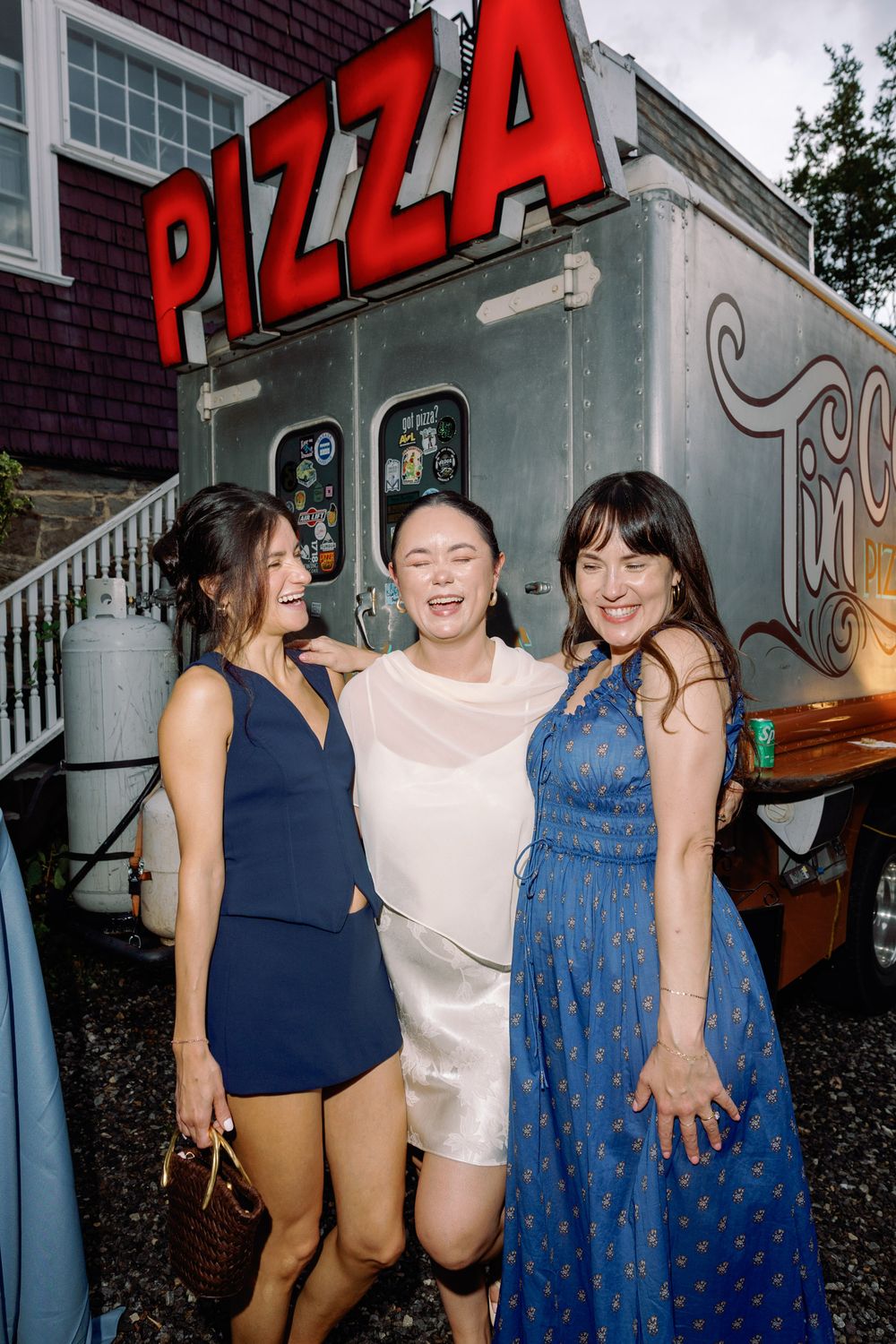 Three women smiling and posing together in front of a pizza food truck at an outdoor event.