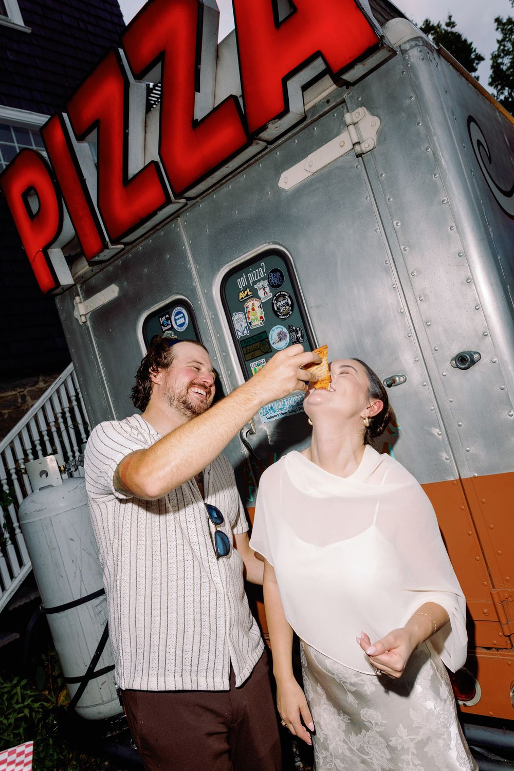 Couple laughing by a pizza food truck, man feeding woman a slice at an outdoor wedding reception.