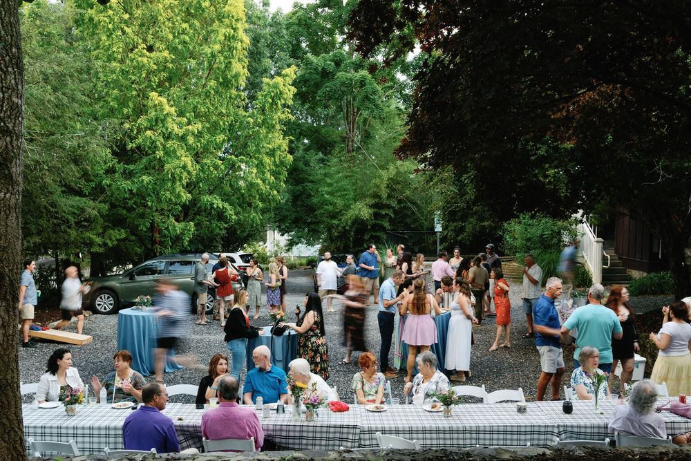 Outdoor garden party with guests mingling and dining at long tables with blue tablecloths surrounded by lush trees.