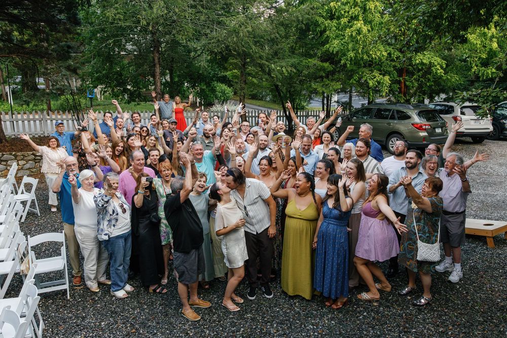 Large group of joyful wedding guests cheering and waving outdoors near white chairs and lush green trees.
