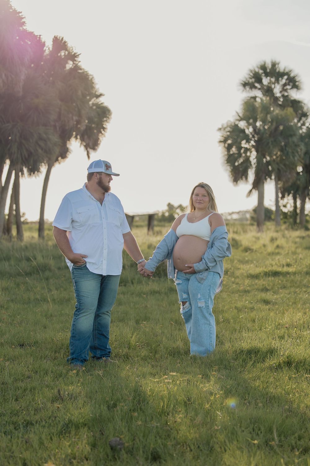 Expecting couple holding hands walking through sunny palm tree field during maternity shoot.