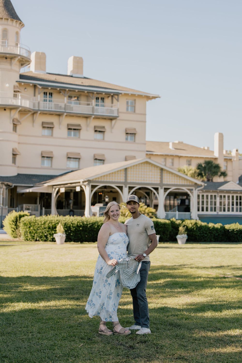 Smiling couple poses on green lawn in front of a grand historic hotel on a sunny day.