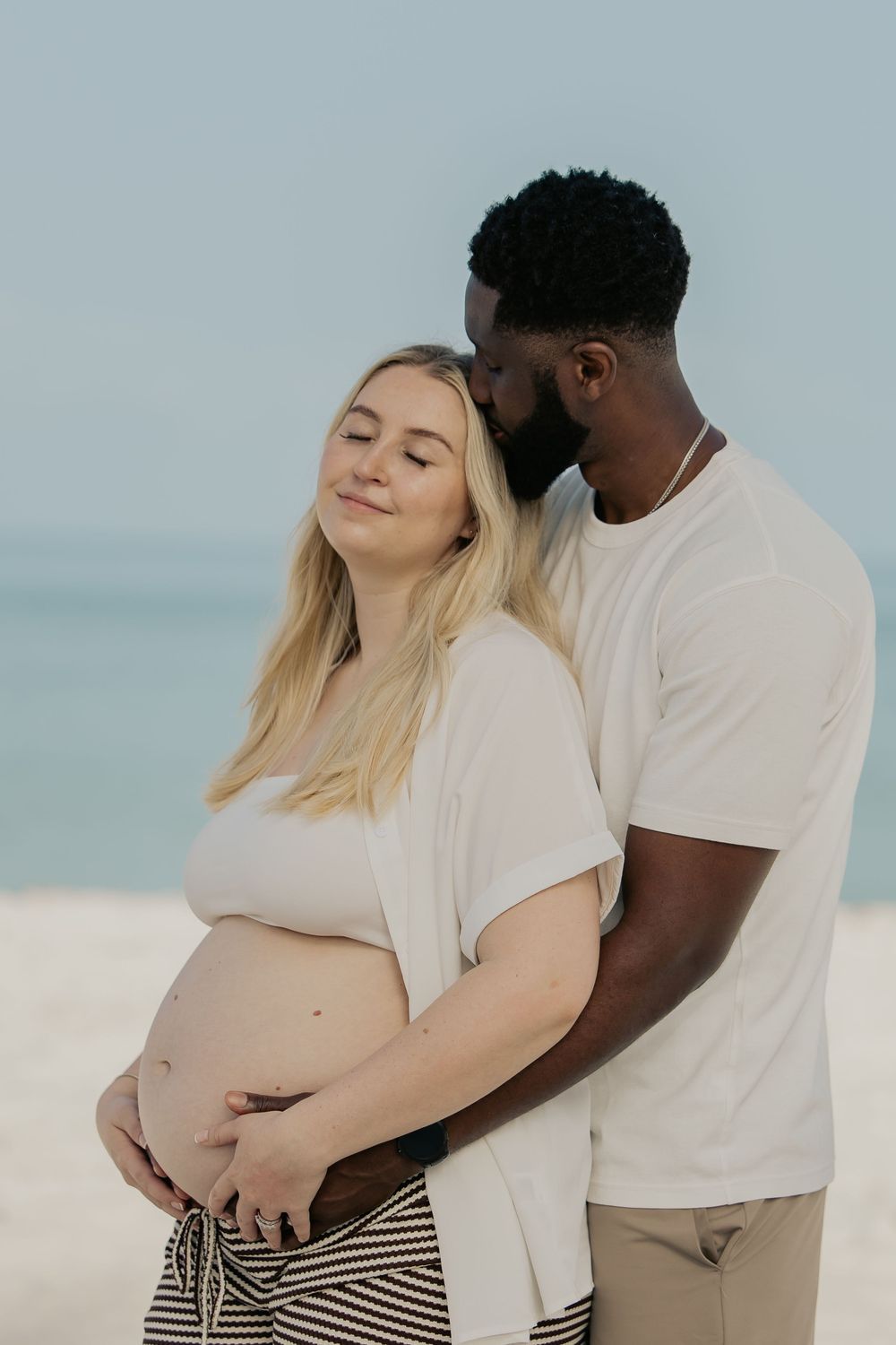Interracial couple sharing tender moment on beach, man embracing pregnant woman from behind.