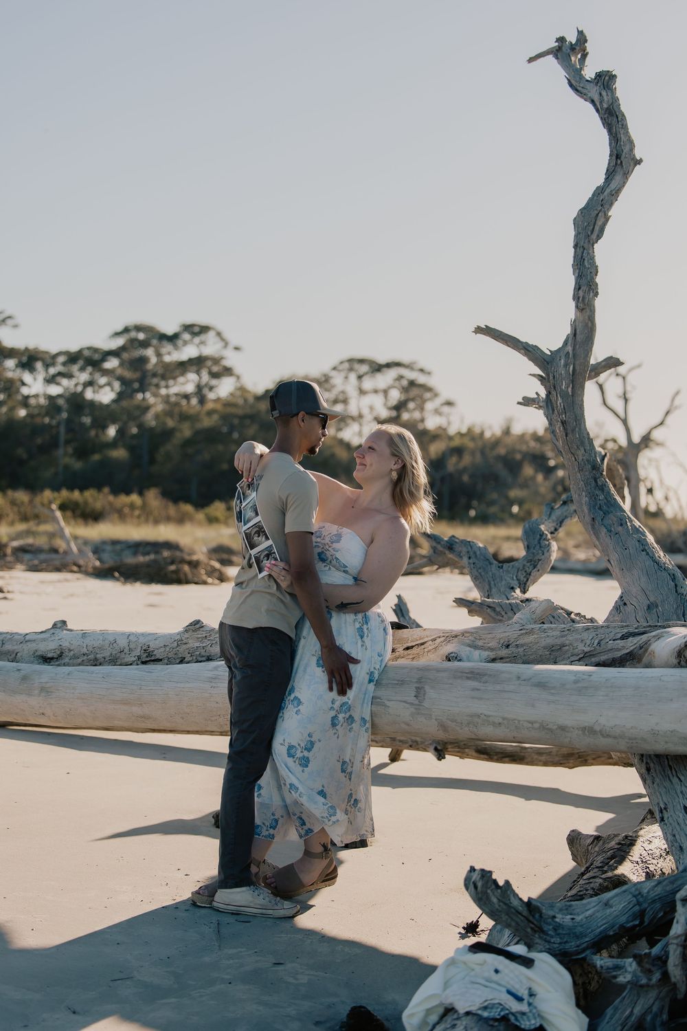 Couple gazes lovingly at each other among driftwood on a sunny beach, holding ultrasound photos.