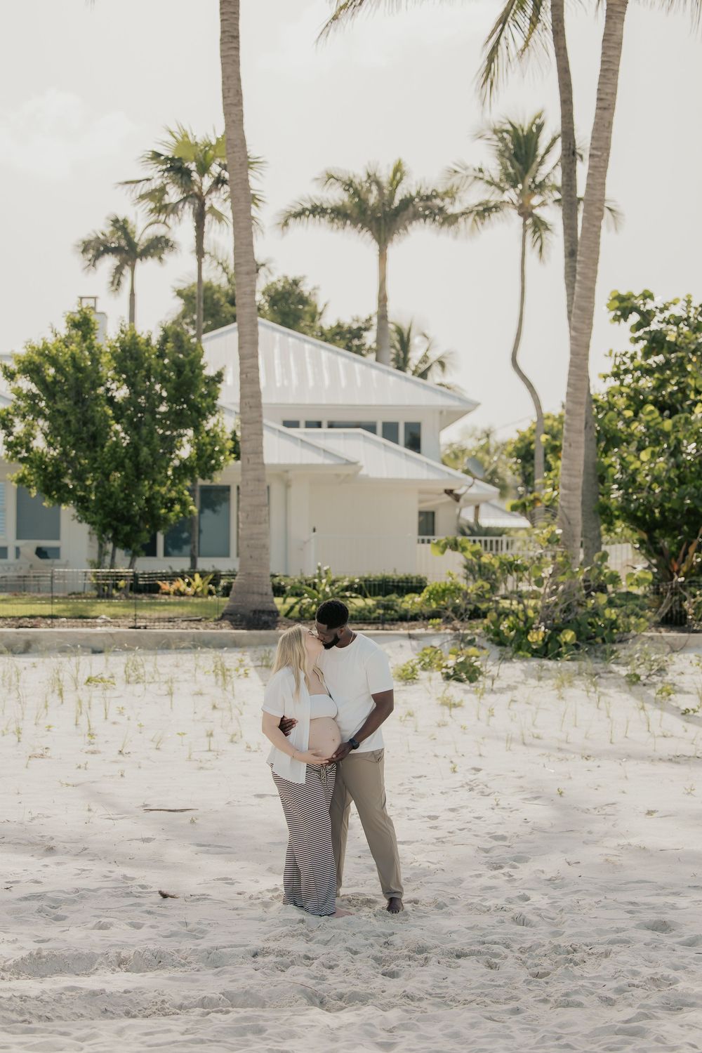 Interracial expecting couple sharing a kiss on sandy beach with palm trees and white house.