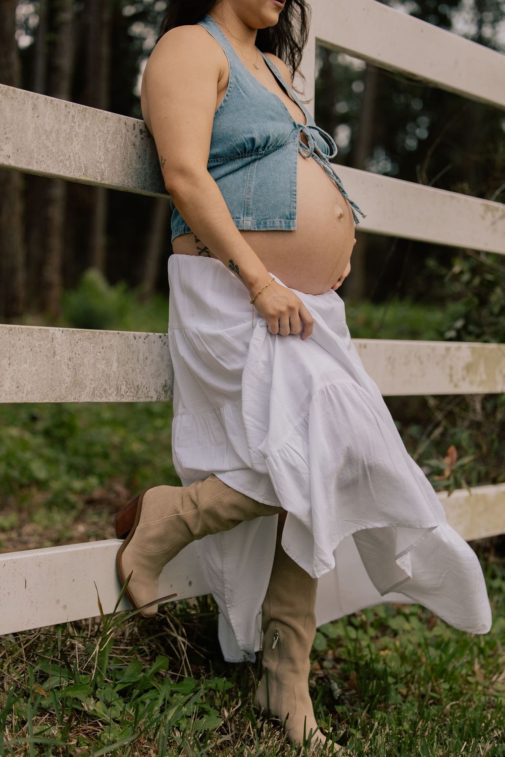 Pregnant woman in denim crop top and white skirt leans against a white wooden fence outdoors.