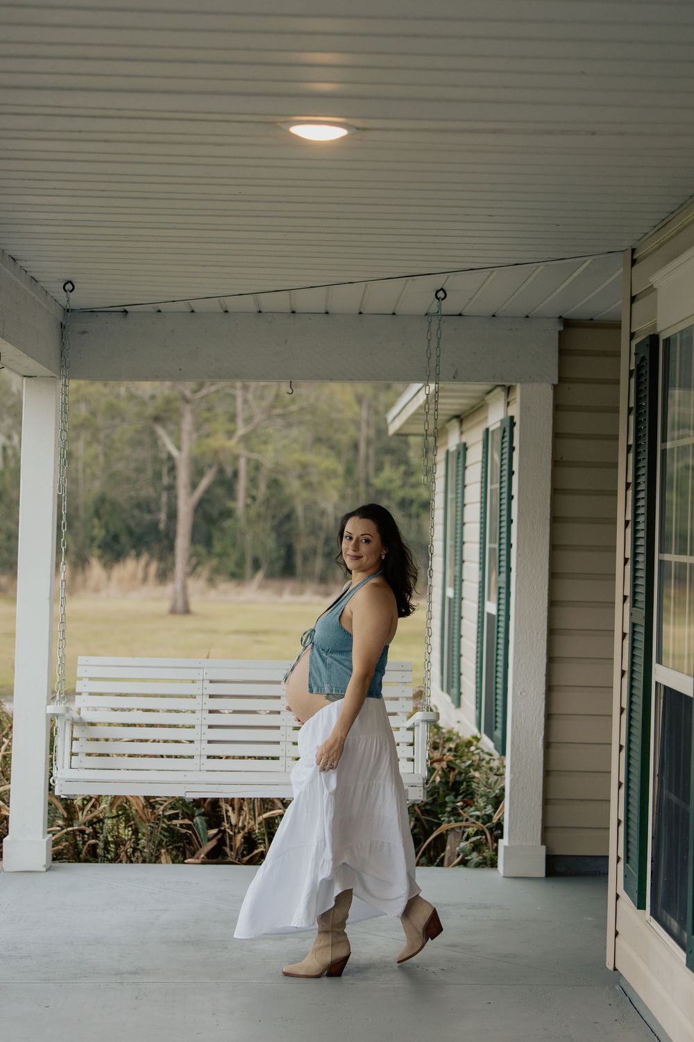 Pregnant woman in blue crop top and white skirt posing on farmhouse porch with cowboy boots.