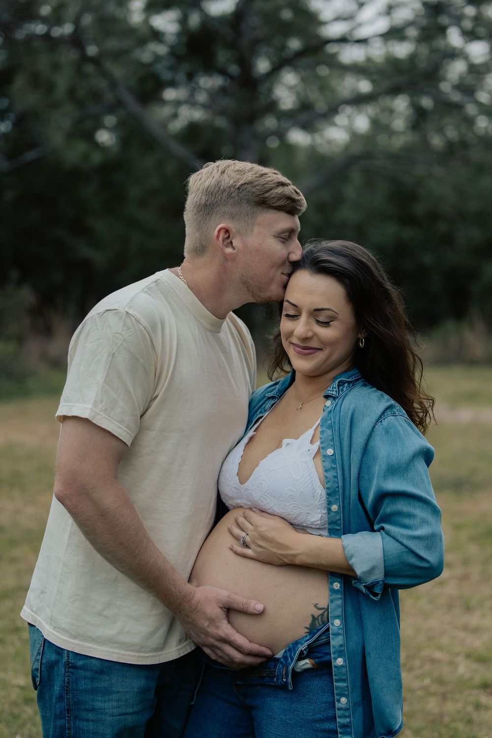 Man kissing pregnant woman's head while cradling her baby bump outdoors in green field.