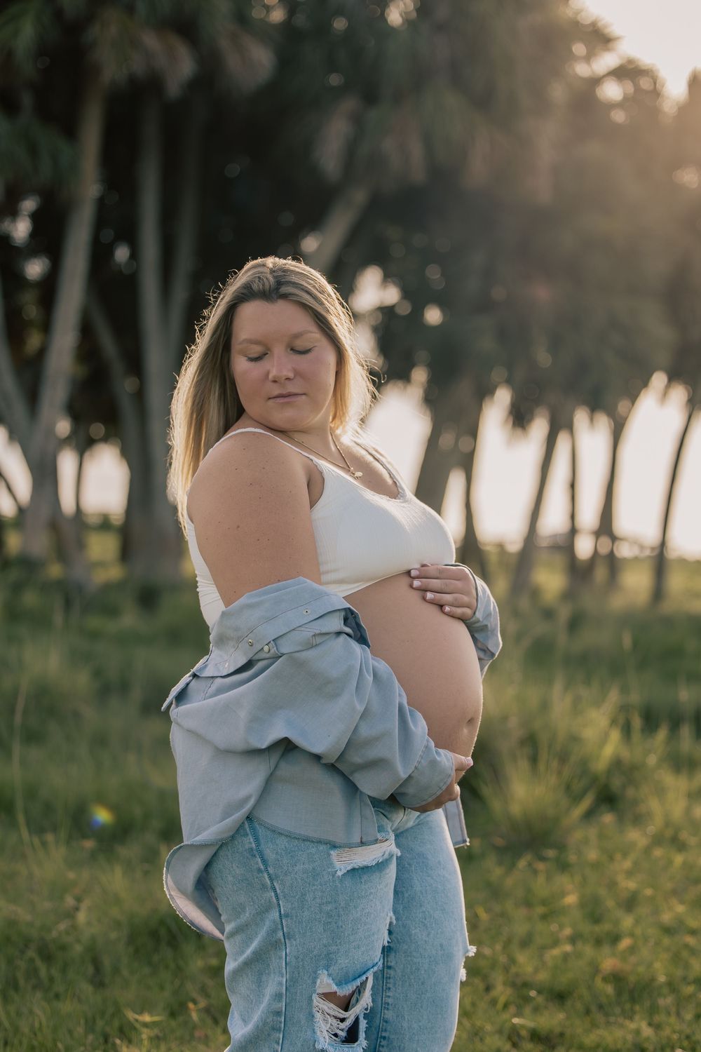 Pregnant blonde woman in denim jacket cradling baby bump during golden hour outdoor session.