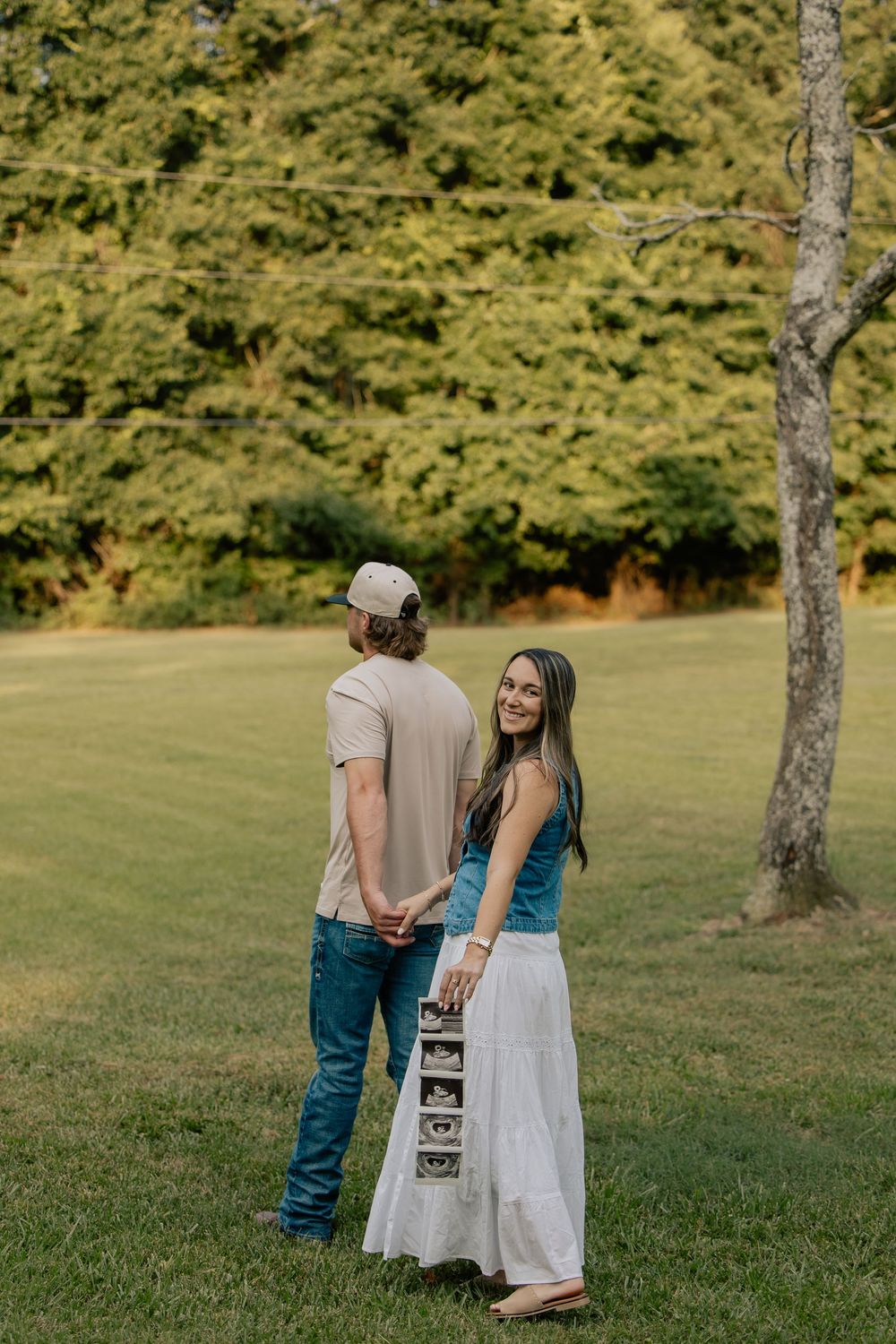 Smiling woman looks back while holding ultrasound strip with partner in a lush green outdoor setting.