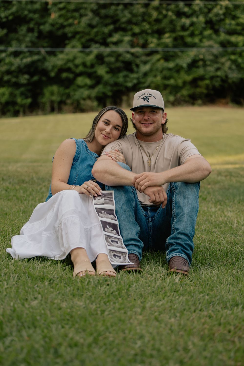 Couple sits on grass smiling, proudly displaying ultrasound photos in a sunny outdoor pregnancy announcement.