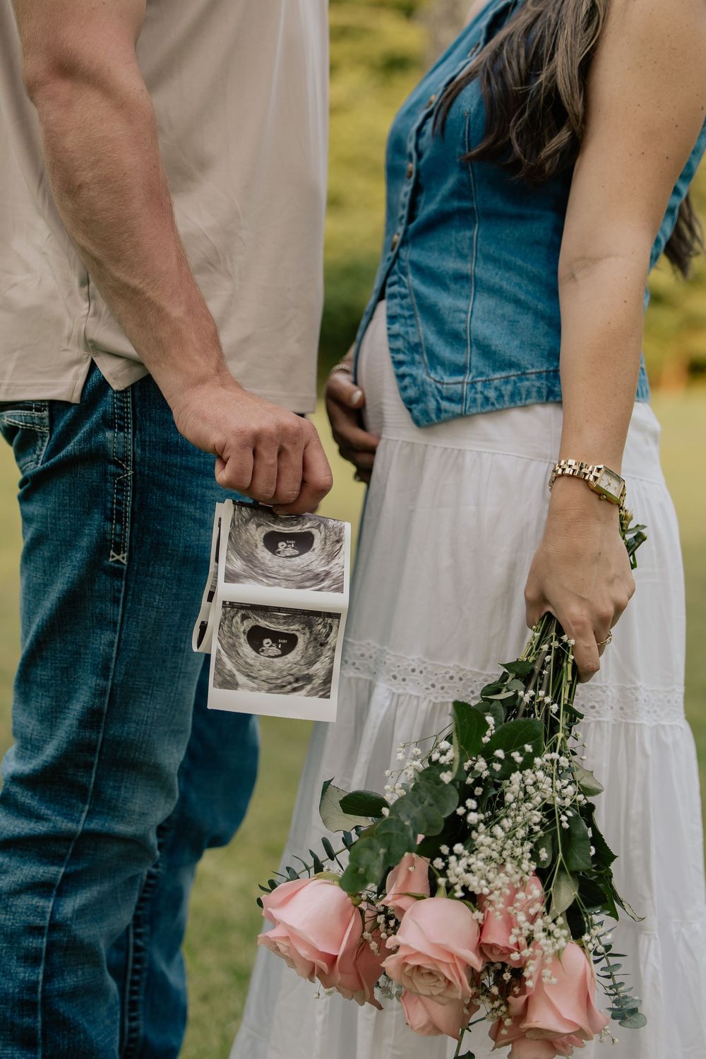 Couple holds ultrasound photos and pink roses bouquet outdoors, announcing pregnancy in a park setting.