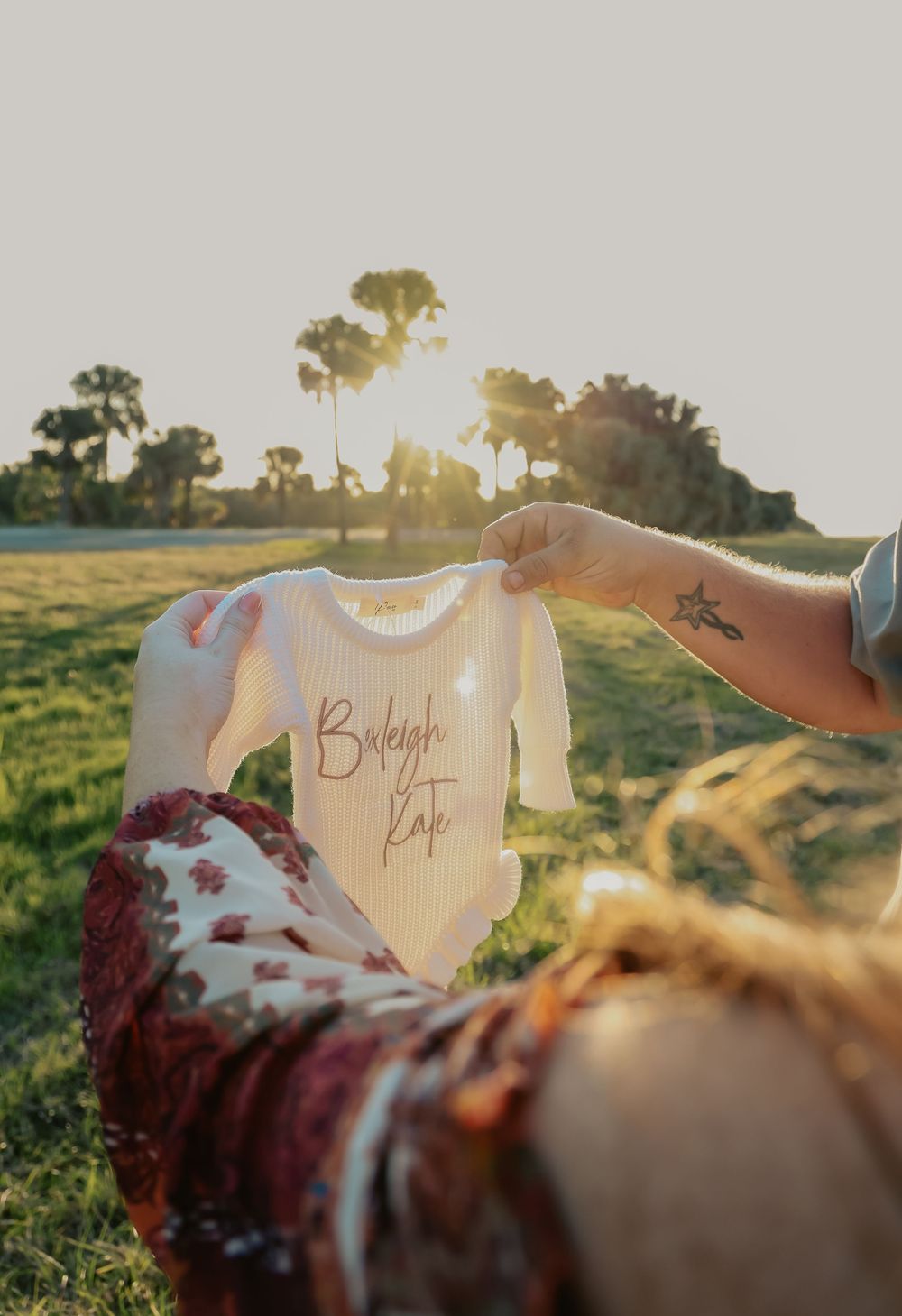 Couple holding personalized baby onesie reading Baleigh Kate during golden hour maternity shoot.