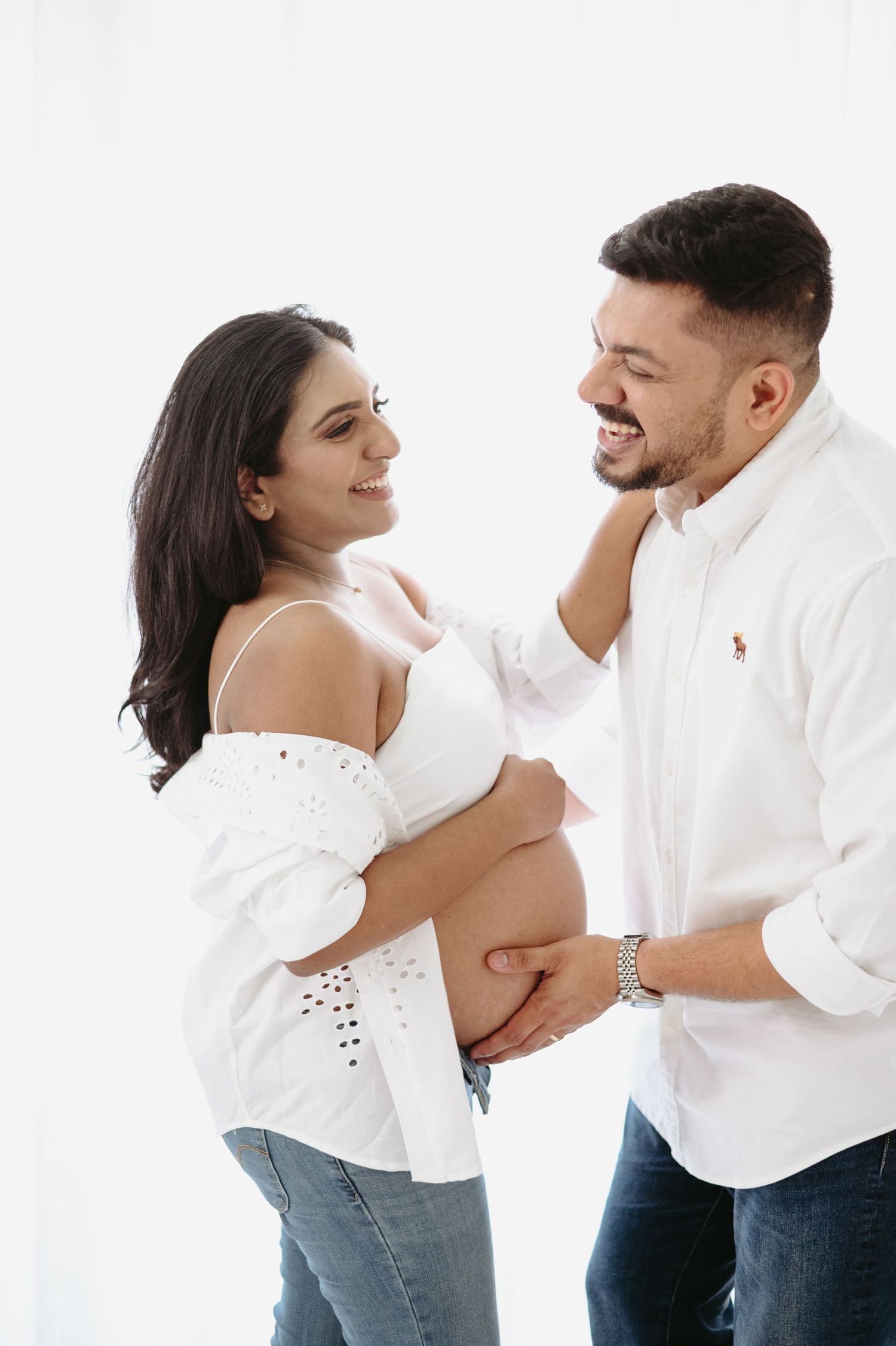 Happy couple in white outfits smiling at each other while man holds pregnant woman's baby bump against white background.