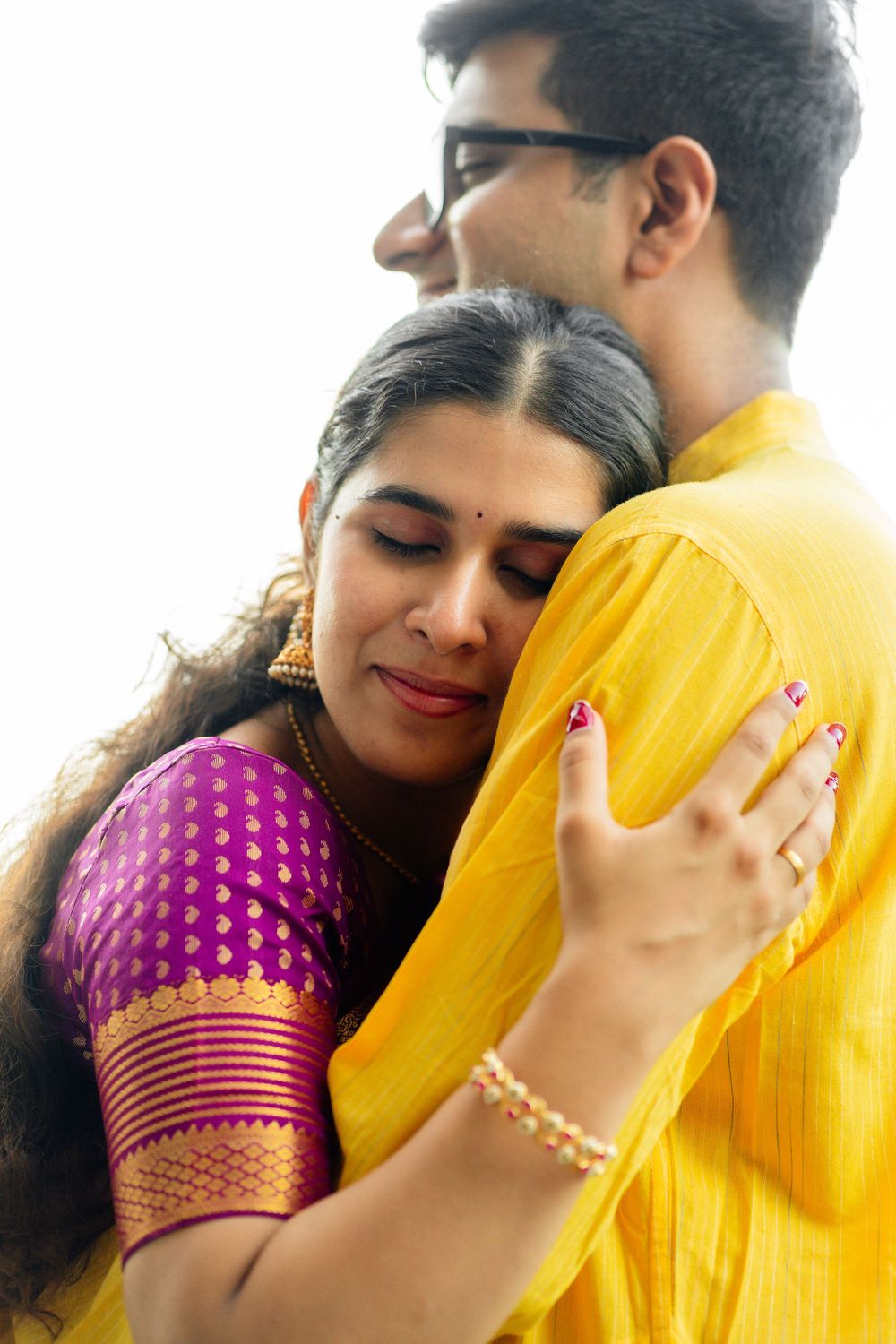 Indian woman in purple and gold saree embracing man in yellow kurta in a tender close-up portrait.