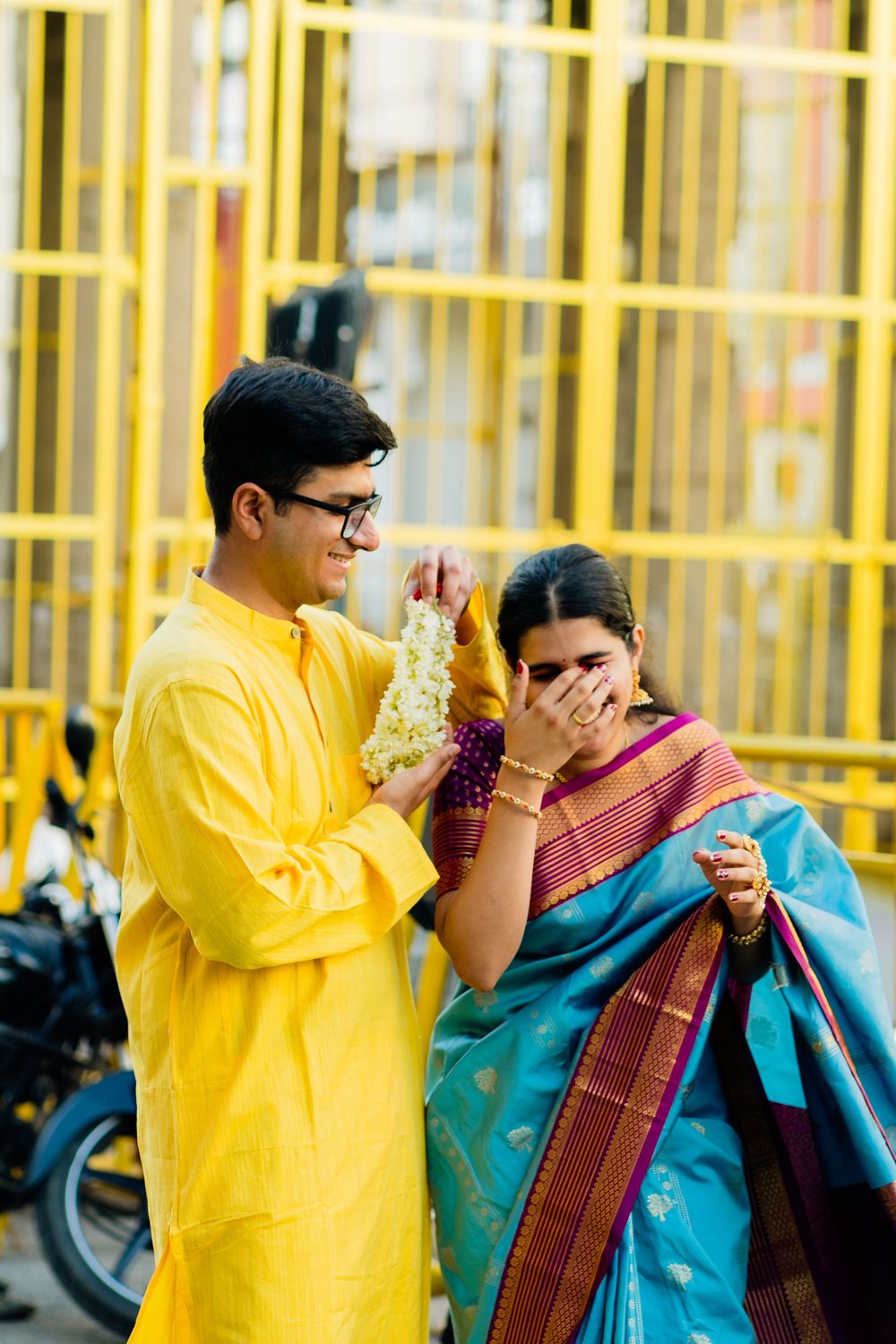 Young Indian couple in traditional attire, man in yellow kurta offers flowers to woman in teal silk saree.