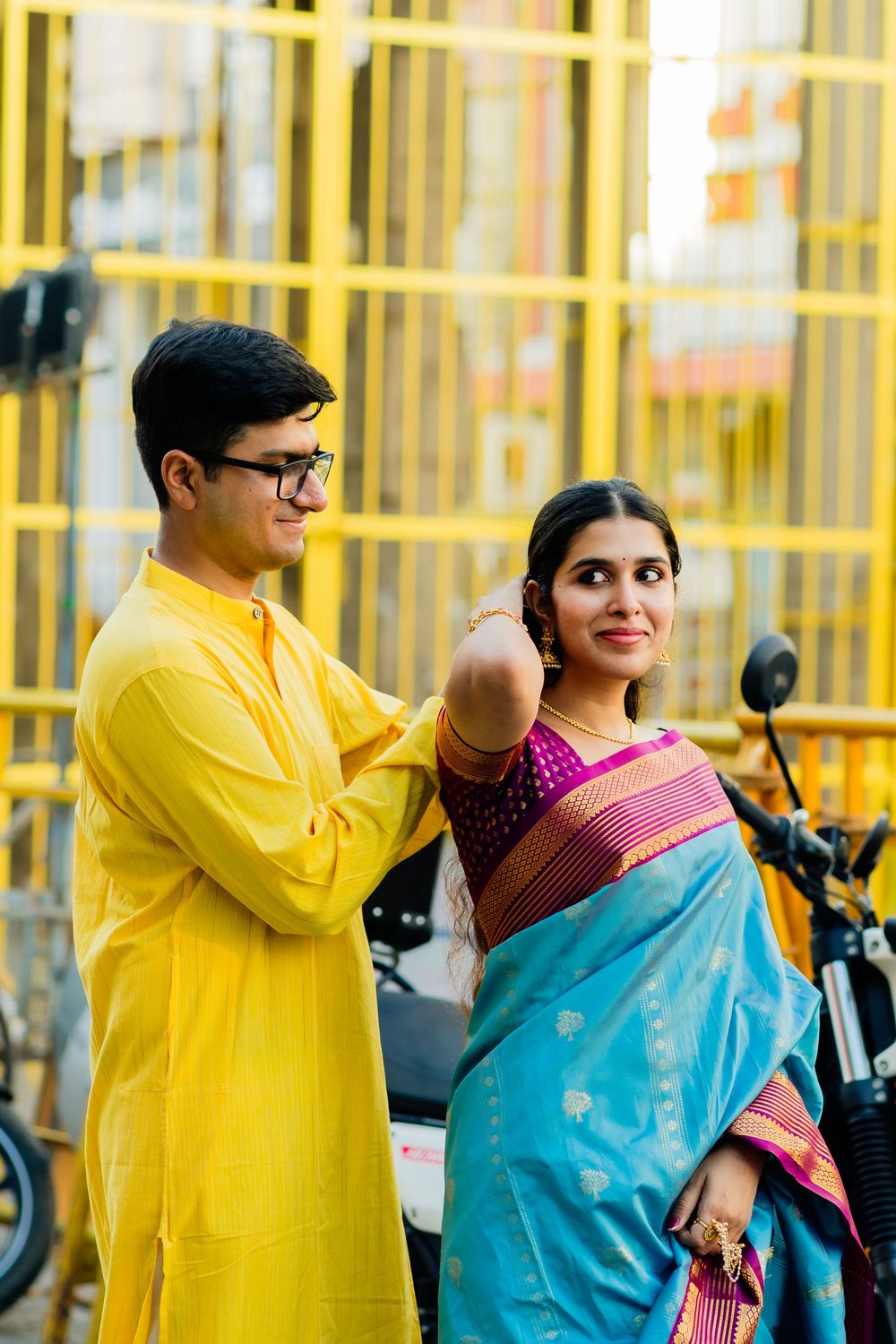 Indian couple in traditional attire posing together near yellow gates with motorcycles.