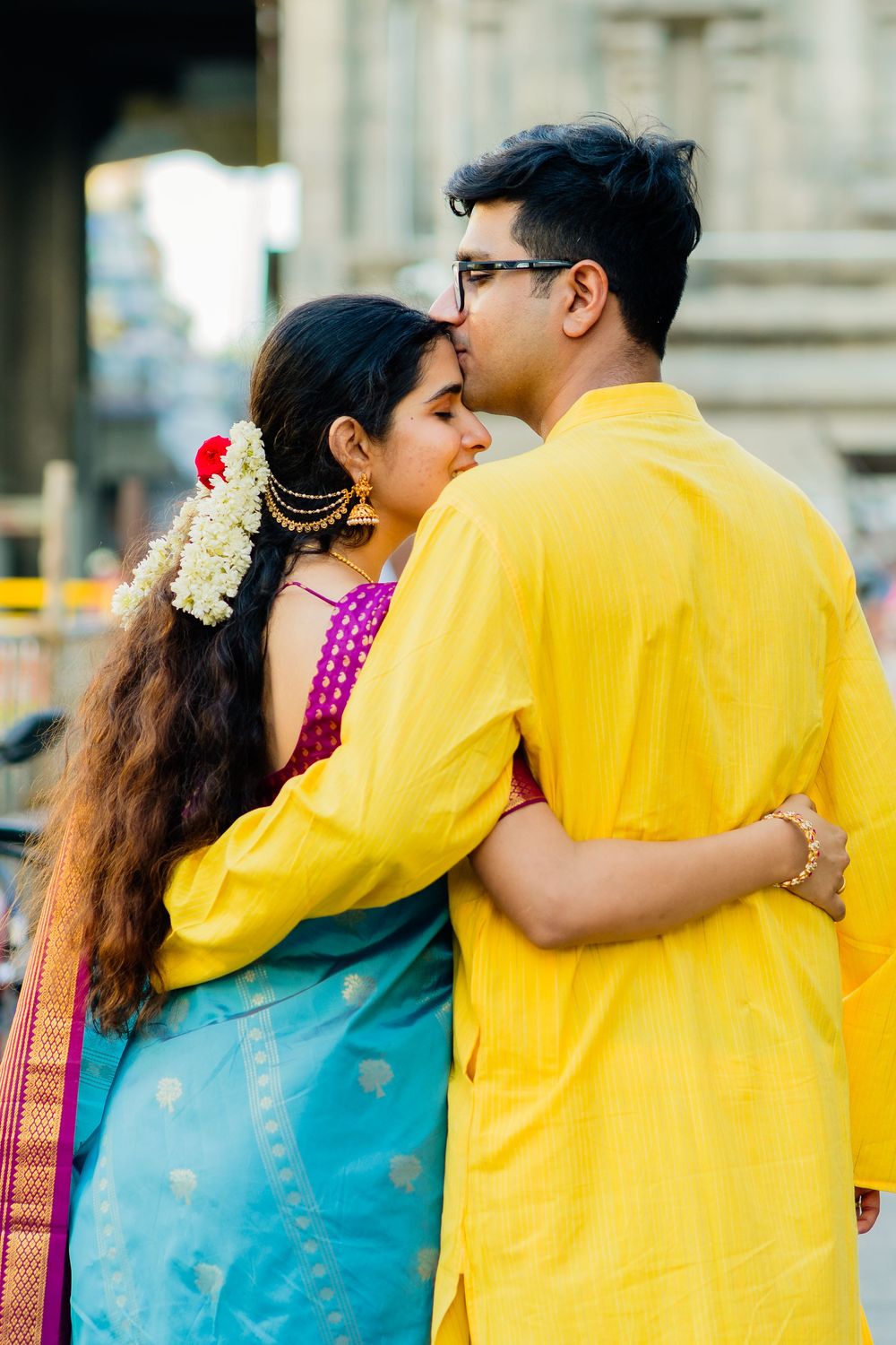 Indian couple in traditional attire embracing, man in yellow kurta kisses woman in blue saree outdoors.