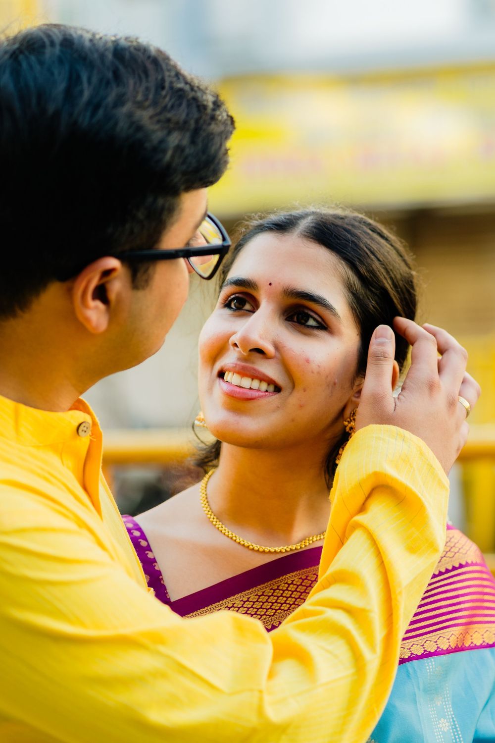 Smiling Indian woman in yellow saree as man gently tucks hair behind her ear outdoors.