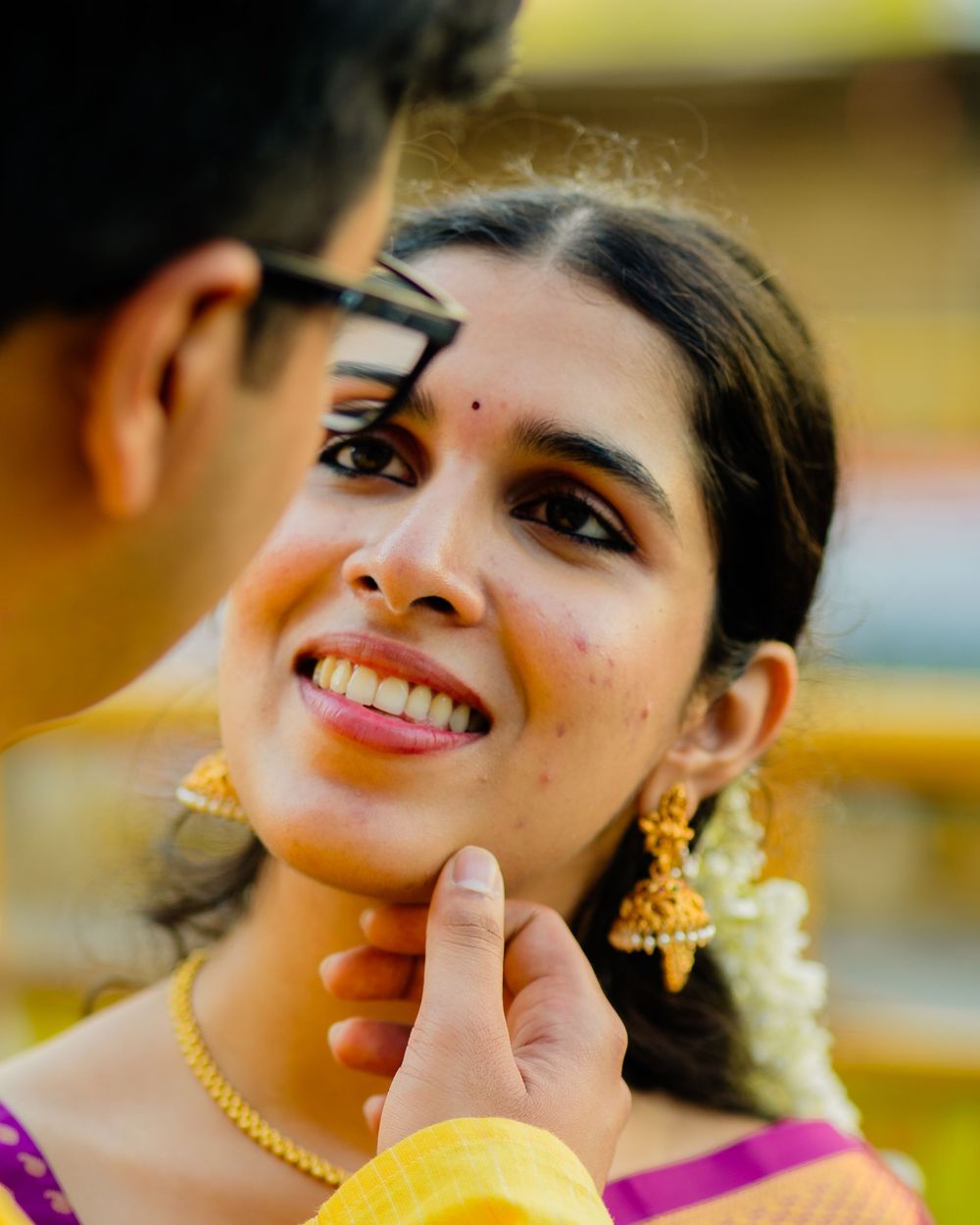 Close-up of smiling Indian bride in gold jewelry being tenderly touched by groom.