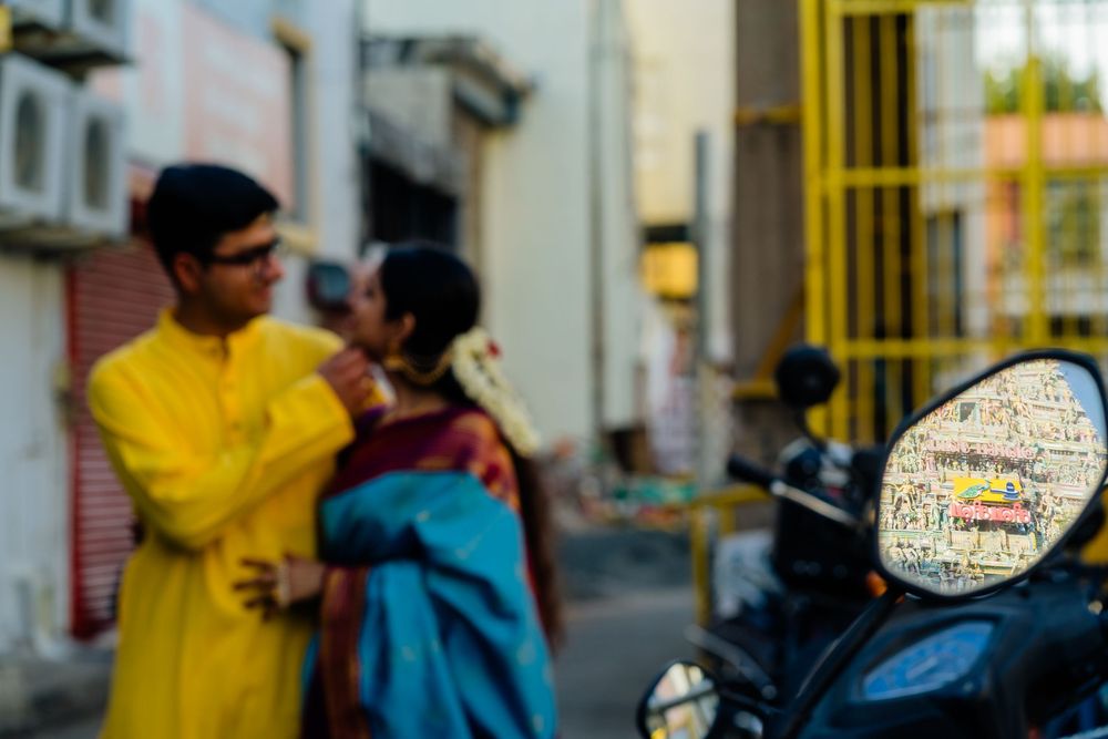 A man in yellow kurta and woman in blue saree stand together on an Indian street, with a scooter mirror in the foreground.