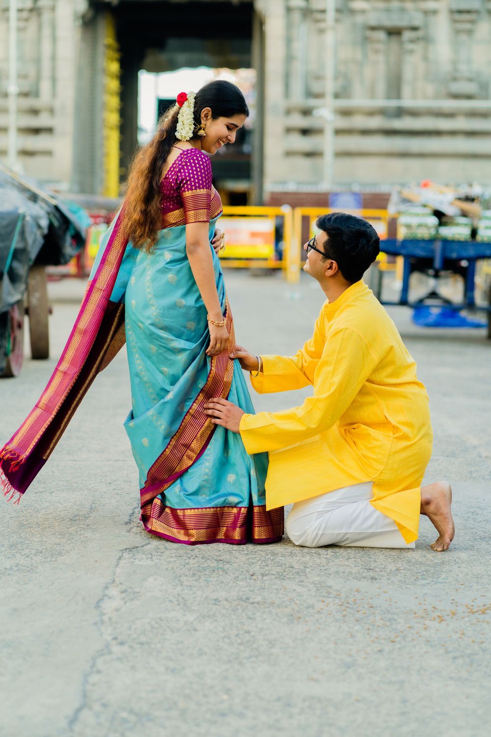 Groom in yellow kurta kneels before bride in teal saree outside a Hindu temple.